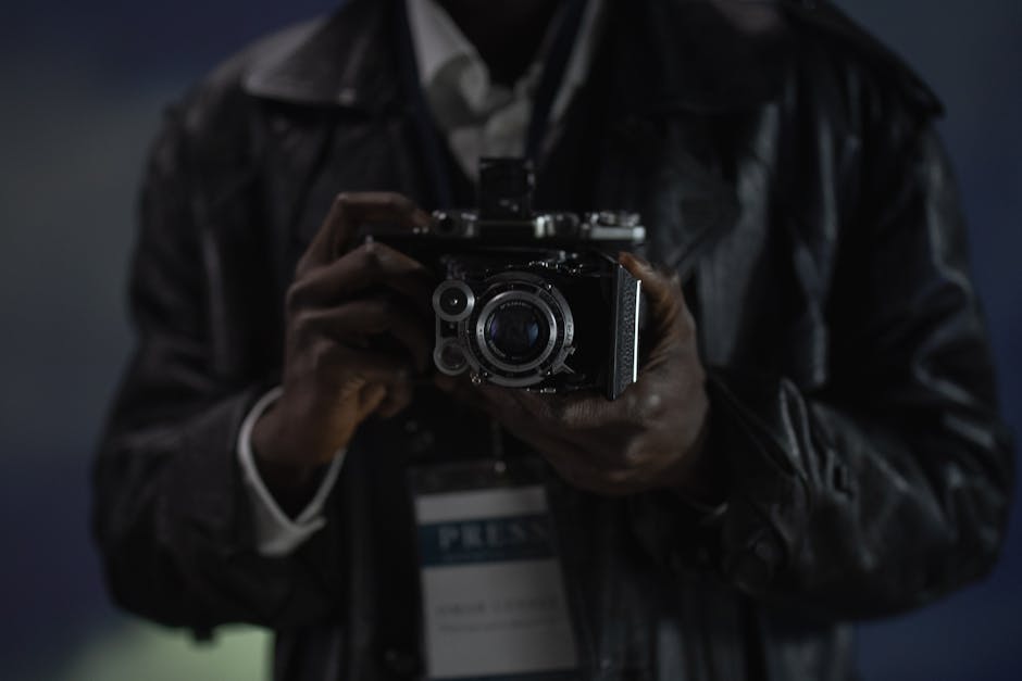 A close-up photograph of a person holding a vintage camera, possibly a reporter.